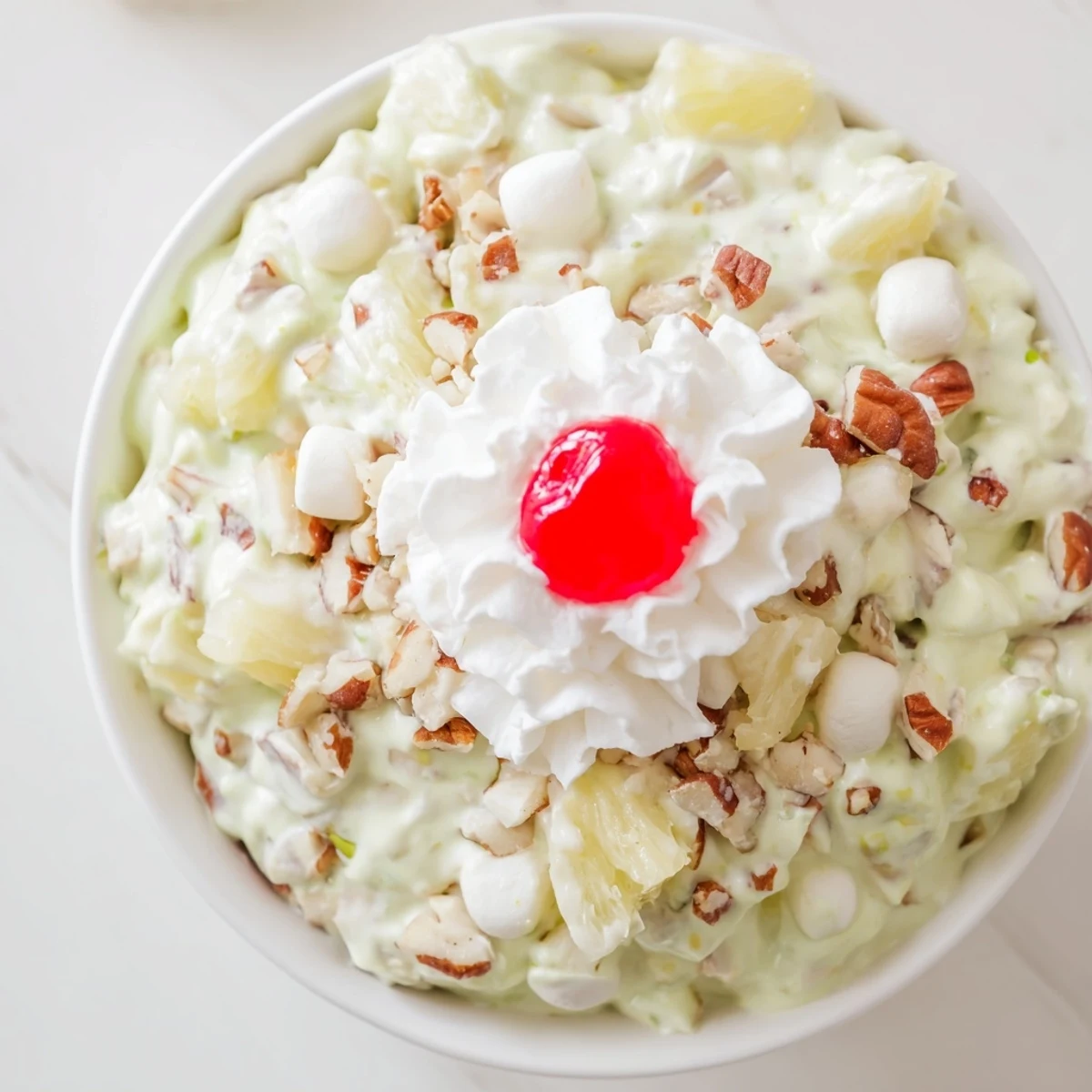 Creamy green Watergate Salad recipe topped with fluffy whipped cream and bright red maraschino cherries in a vintage glass bowl